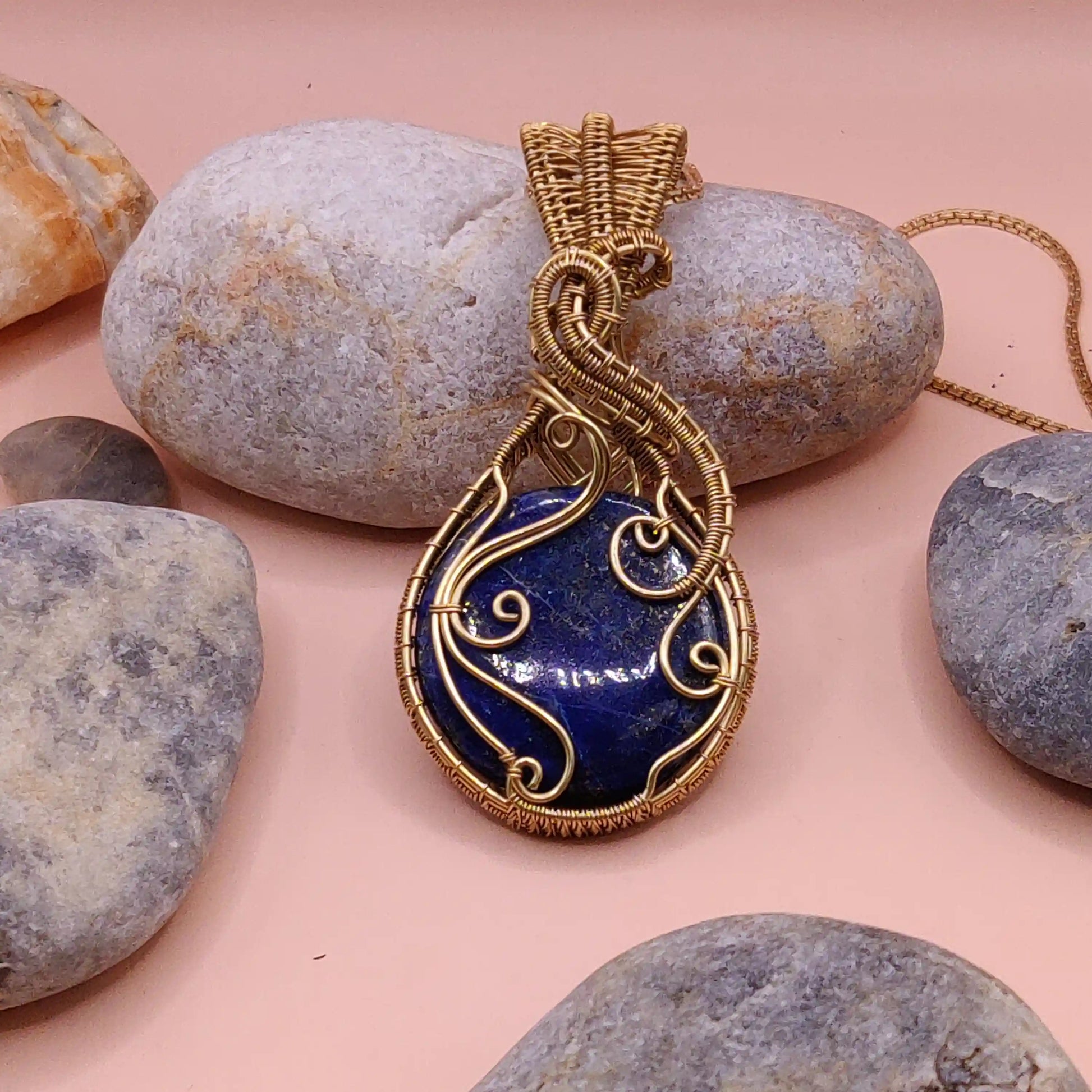 Gold brass-wrapped blue lapis lazuli stone pendant on a pink surface with stones around