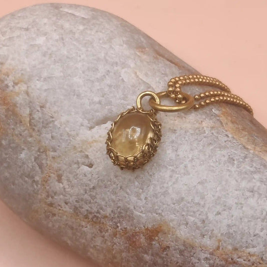 A brass chain necklace with a small oval pendant featuring a natural Citrine, draped over a smooth gray decorative rock on a soft peach-pink background.